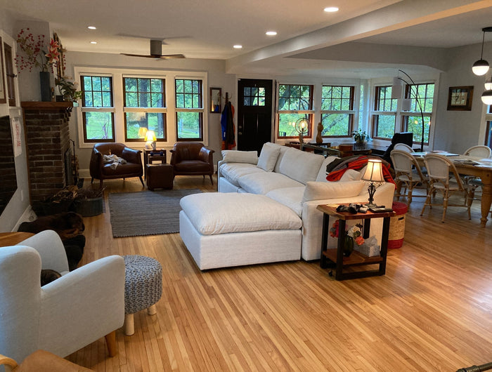 A cozy living room with a large white sectional sofa, wooden floors, and a brick fireplace. There are armchairs, a dining table, bookshelves, and multiple windows letting in natural light.