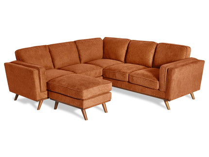 A spacious loft with brick walls, large windows, and concrete floors features an orange sectional sofa, a circular white rug, a wooden desk, a chair, a plant, and a set of shelves in the background.