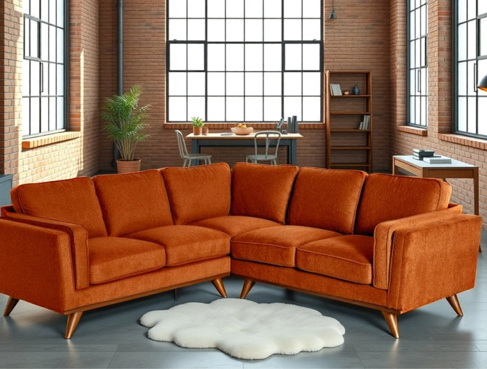 A spacious loft with brick walls, large windows, and concrete floors features an orange sectional sofa, a circular white rug, a wooden desk, a chair, a plant, and a set of shelves in the background.