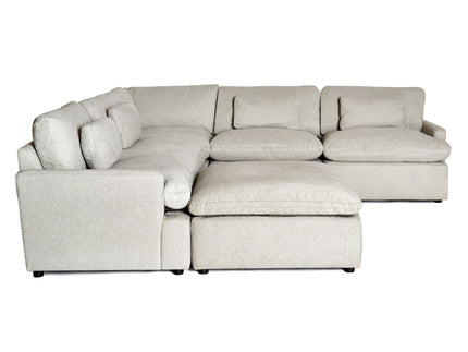 A cozy living room with a large gray sectional sofa, a round wooden coffee table, a patterned area rug, and an armchair with a mustard cushion. The space is bright with large windows showing greenery outside.