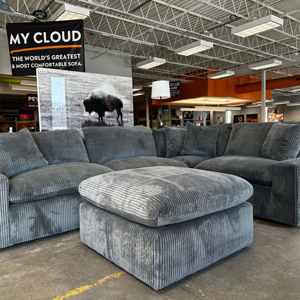 A large, plush gray sectional sofa with a matching ottoman is displayed in a showroom. A sign above reads, MY CLOUD: The Worlds Greatest & Most Comfortable Sofa. A bison image is in the background.