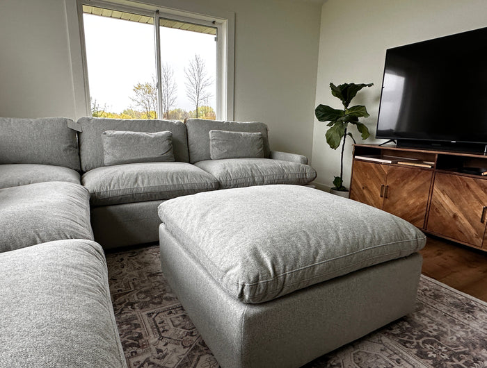 A cozy living room with a large gray sectional sofa and ottoman, a flat-screen TV on a wooden stand, a patterned rug, a fiddle leaf fig plant, and a window with a view of trees outside.