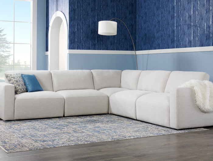 Modern living room with a large, light gray sectional sofa, a blue patterned accent wall, and a standing floor lamp. A dark blue and white rug lies on a dark wood floor. A window lets in natural light.