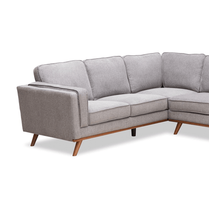 The Maddox LSF is a gray, L-shaped sofa with wooden legs and plush backs, displayed on a white background. It features a modern design and clean lines inspired by mid-century aesthetics.