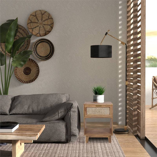 A cozy living room showcases a gray sofa, the Bedside Table 1 Drawer holding a plant, and a black and gold lamp. Woven cane panels accent the walls, and wooden blinds on the large window allow natural light to fill the space.