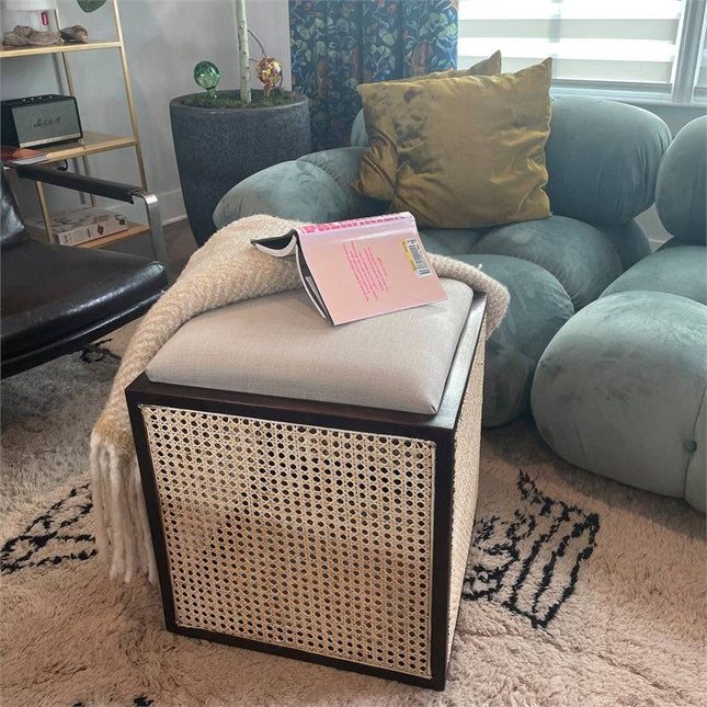 A cozy living room includes a green plush chair, an Upholstered Stool With Cane topped with a book and blanket, a patterned rug, and a large window with blinds. Decorative accents like a plant, lamp, and colorful pillows make it a multifunctional space.