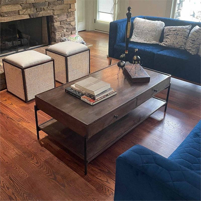 A cozy living room features a wooden coffee table with three books and decor. A navy sofa with cushions sits beside two Upholstered Stools With Cane. A handcrafted Cane Cube Ottoman adds a multifunctional accent, while the stone fireplace complements the hardwood floor in the background.