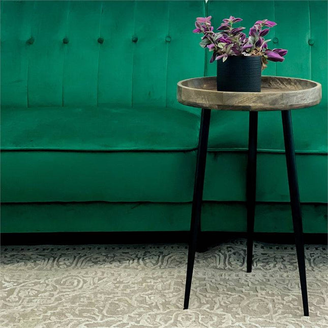 A small Mango Bowl Side Table with a mid-century design and black legs holds a purple-leafed potted plant. The mango wood piece sits on a patterned rug in front of a green upholstered sofa.