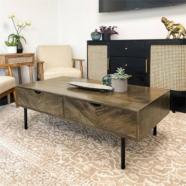 A modern living room showcases a Mango 4 Drawer Media Unit topped with a decorative tray and small plant. It is surrounded by two beige armchairs and a black cabinet with woven panels, all set on a textured beige rug.