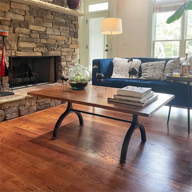 A cozy living room features an Acacia Foundry Coffee Table on a wooden floor, showcasing books and decorative bird sculptures with vintage-inspired style. Behind it is a stone fireplace, a blue sofa with cushions, a glass terrarium, and a floor lamp near the window.