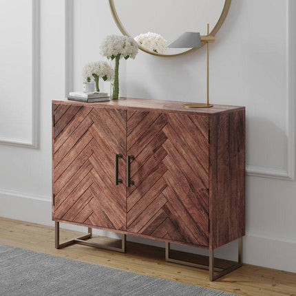 An Acacia Wooden 2 Door Cabinet with Iron Leg is set against a white wall. It features a modern lamp, two books, and white flowers in a vase on top. A round mirror elegantly reflects the room above it, while a gray rug adorns the wooden floor below.
