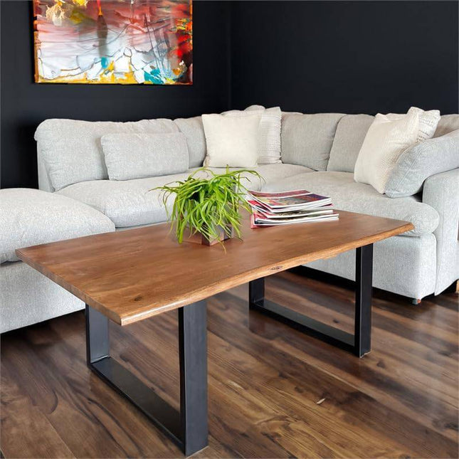 A modern living room showcases a gray sectional with white pillows, the Live Edge Coffee Table 1 Tier featuring black metal legs, magazines stacked on top, and a green plant. A colorful abstract painting adorns the dark wall, complementing the wooden floor.