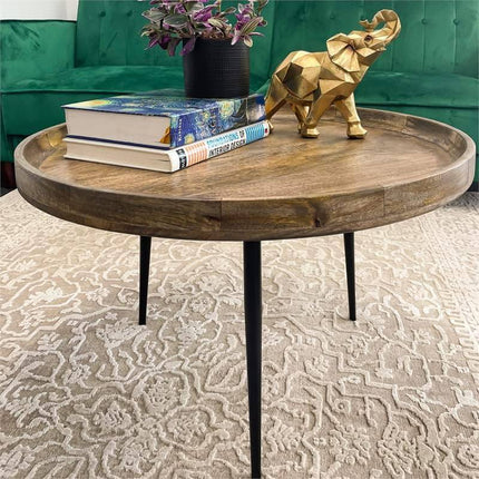 The Mango Bowl Coffee Table with black legs showcases a stack of books, a gold elephant figurine, and a small plant. This mid-century piece sits on a patterned beige rug in front of a green sofa, seamlessly combining classic charm and modern ethics.