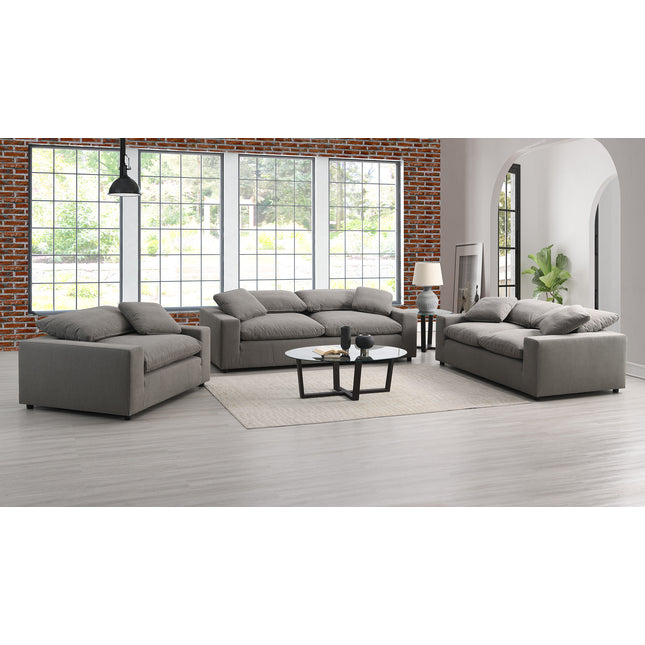 A modern living room with a Peyton Classic Sofa around a round coffee table. Large windows, a brick wall accent, light wood flooring, and a potted plant by the archway add to the décor, ideal for contemporary spaces.