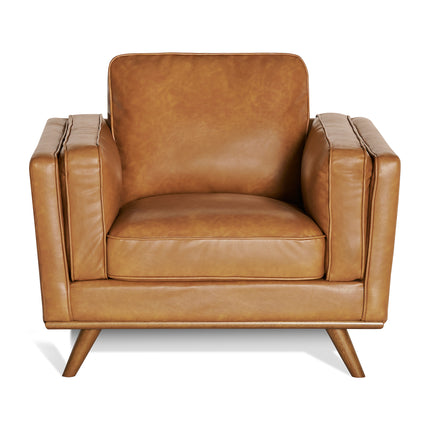 The Maddox Chair, a brown leather armchair with a mid-century modern boxy design, is centered in the image. It showcases plush cushions, wide armrests, a squared back, and tapered wooden legs on a plain white background.