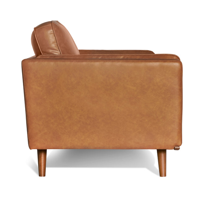 The Maddox Chair features a mid-century modern style with light brown leather upholstery, plush cushions, and wooden legs. Its boxy design includes a subtle sheen on the material.