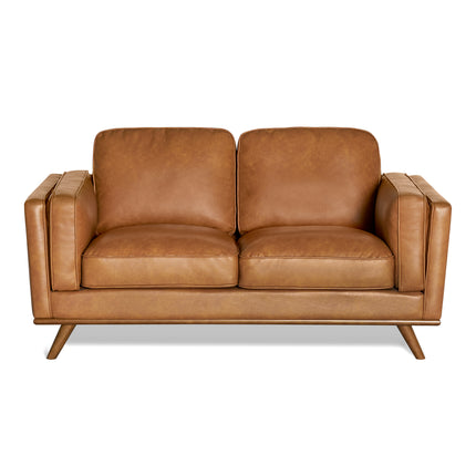 The Maddox Loveseat, a durable mid-century modern-style piece with brown leather, boxy cushions, and wooden tapered legs, fits perfectly in an empty white space.