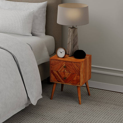 A modern bedroom features the MCM Bedside Table made from solid mango wood with tapered legs. It holds a marble lamp with a beige shade, a small white clock, and a black circular object. The chic setup is completed by the beds white bedding on a geometric-patterned beige carpet.