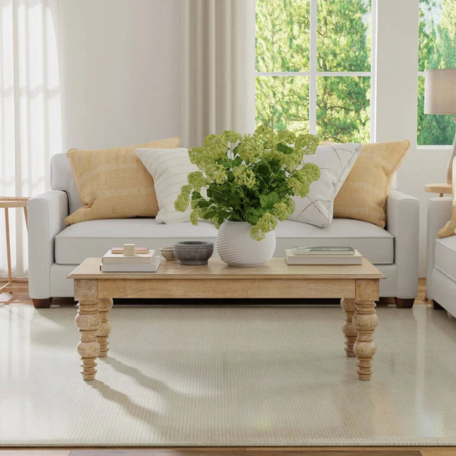 A bright living room features a white sofa with beige and white cushions. The French Country Coffee Table with a distressed natural finish sits on a light carpet, adorned with a potted plant and books. Large windows reveal green foliage outside, complemented by a solid mango wood dining table.