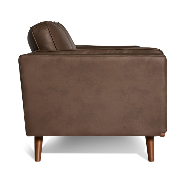 Side view of a brown leather armchair with a boxy mid-century modern design, plush cushions, and four short tapered wooden legs. Lighting highlights the leather texture and clean lines of this Maddox Sofa piece.