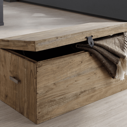 The Rustic Wooden Chest, with simple metal handles and a partly open lid, is featured in a minimalist room. A folded patterned blanket with tassels drapes over the edge of the chest, which sits on a light wooden floor.
