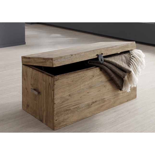 The Rustic Wooden Chest, with simple metal handles and a partly open lid, is featured in a minimalist room. A folded patterned blanket with tassels drapes over the edge of the chest, which sits on a light wooden floor.