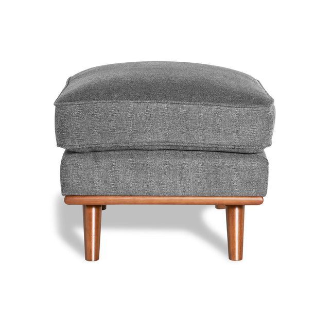 The gray upholstered Maddox Ottoman features a plush cushion atop four wooden legs, blending sleek mid-century design with the softness of fabric and warmth of wood. Bright lighting casts shadows beneath, making it ideal for any modern living space.
