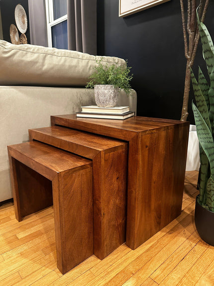 Beside a beige sofa, a set of three Acacia Nesting Tables is arranged. The top table holds stacked books and a potted plant. Ideal for small spaces, the room features a wooden floor with a tall plant to the right.