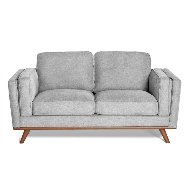 The Maddox Loveseat features light gray upholstery with plush cushions, a wooden base with angled legs, showcasing mid-century modern design. It boasts clean lines, minimalist aesthetics, and durable construction.