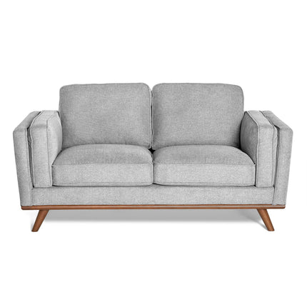 The Maddox Loveseat features light gray upholstery with plush cushions, a wooden base with angled legs, showcasing mid-century modern design. It boasts clean lines, minimalist aesthetics, and durable construction.