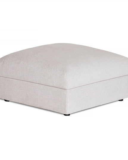 The Lugano Storage Ottoman, a square, beige piece with a textured fabric and smooth cushioned top, rests on a white background. Its sleek modern design is accented by small black feet that are subtly visible underneath.