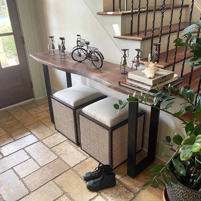 A hallway showcases the Live Edge Console adorned with artistic bicycle sculptures, books, and a shell. Below sit two uniquely styled storage benches. Nearby are black shoes and a plant on eco-friendly tile flooring, with stairs featuring a black railing in the background.