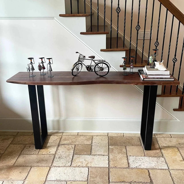 The Live Edge Console, made from acacia wood with black metal legs, features metal bicycle sculptures and figures. On a stone-tiled floor near a staircase with black wrought iron railings, its adorned with books and an eco-friendly white decor item.