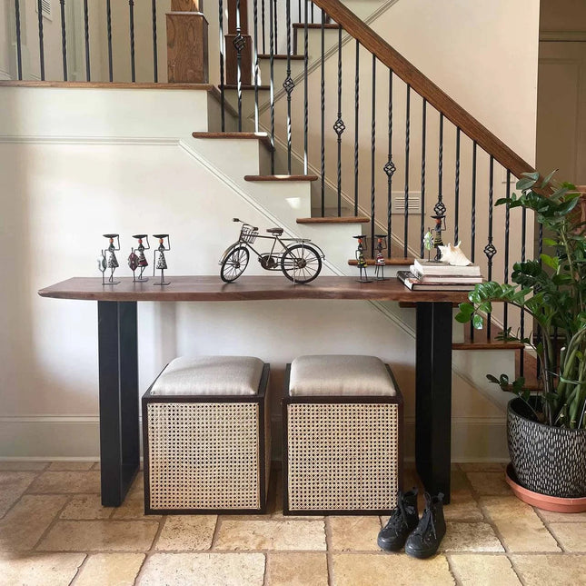 An entryway featuring a Live Edge Console with unique styles, adorned with bicycle sculptures and decor. Two cushioned storage cubes rest below. Wrought iron staircase railings and a potted eco-friendly plant are in the background, with shoes nearby.