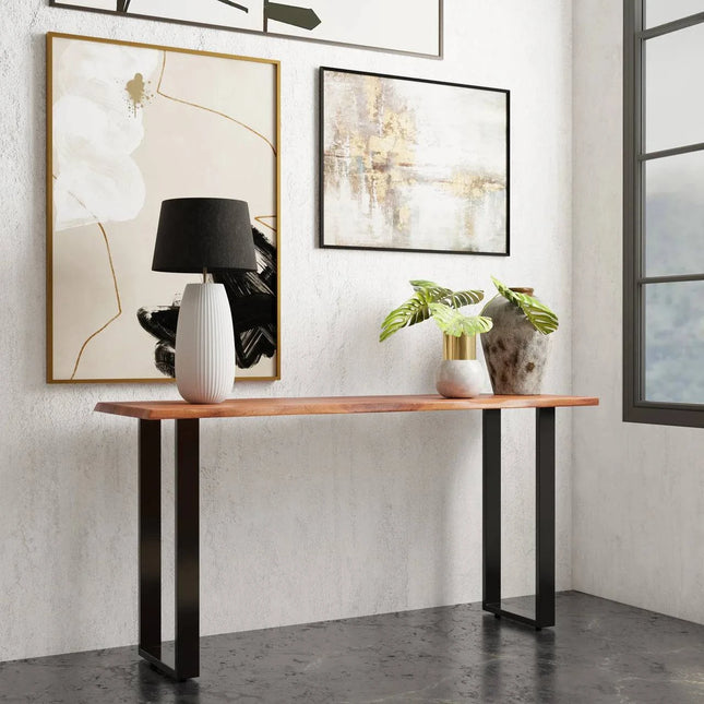 A modern room features the Live Edge Console, a wooden table with a black metal frame and live edge acacia wood. A white lamp with a black shade and a plant sit atop it. Behind are three abstract paintings on a textured white wall, while an eco-friendly blurred outdoor view is visible through the window on the right.