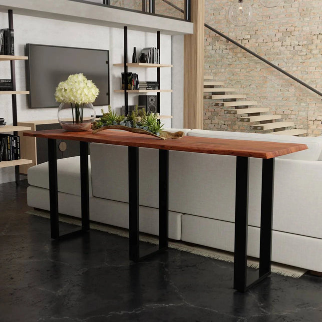 A stylish living room showcases the Live Edge Console, featuring a live edge acacia wood top and black metal legs, adorned with a glass vase of white flowers and a decor item. In the background, theres a white couch, exposed brick wall, bookshelves, and staircase.