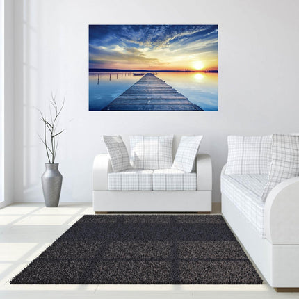 A modern living room showcases a white sofa, gray cushions, and a large vase with tall branches. Sunset Dock, a museum-quality artwork of a vibrant wooden pier at sunset, adorns the wall. Sunlight streams through tall windows onto the tempered glass coffee table and dark rug.