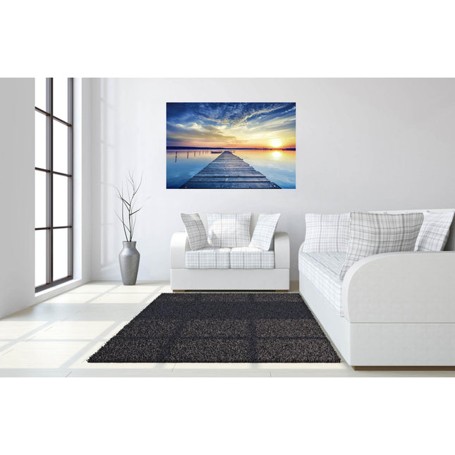 A modern living room showcases a white sofa, gray cushions, and a large vase with tall branches. Sunset Dock, a museum-quality artwork of a vibrant wooden pier at sunset, adorns the wall. Sunlight streams through tall windows onto the tempered glass coffee table and dark rug.