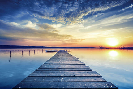 The Sunset Dock extends over a tranquil lake, reflecting the sunsets vibrant hues like museum-quality art. The sky merges deep blues with warm oranges and scattered clouds, while a small boat moored beside the dock adds to the serene ambiance.