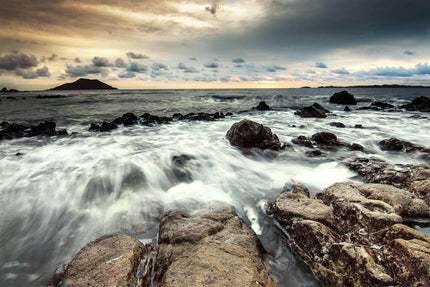 Waves crash against the rocky coast under a dramatic, cloudy sunset, creating a scene reminiscent of museum-quality artwork. The island silhouette on the horizon and the blend of orange and gray hues leave a lasting impression, perfectly capturing the essence of Shoreline.