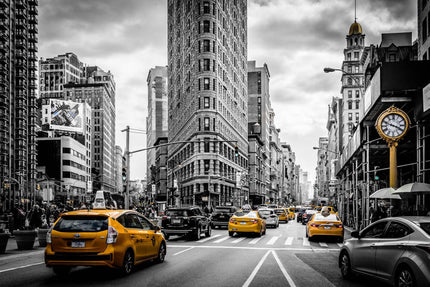 The NYC artwork depicts a lively city street with yellow taxis cruising by New Yorks iconic Flatiron Building. A flurry of cars and pedestrians evokes the feel of a museum piece in mostly black-and-white, highlighting only the taxis and a sidewalk clock in color.