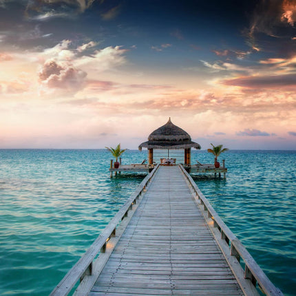 The Ocean Hut, a quaint thatched-roof structure, sits at the end of a wooden pier extending into the serene ocean, surrounded by palm plants. Dramatic clouds filled with orange, pink, and purple hues at sunset create a museum-quality scene under floating tempered glass.