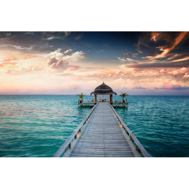 The Ocean Hut, a quaint thatched-roof structure, sits at the end of a wooden pier extending into the serene ocean, surrounded by palm plants. Dramatic clouds filled with orange, pink, and purple hues at sunset create a museum-quality scene under floating tempered glass.