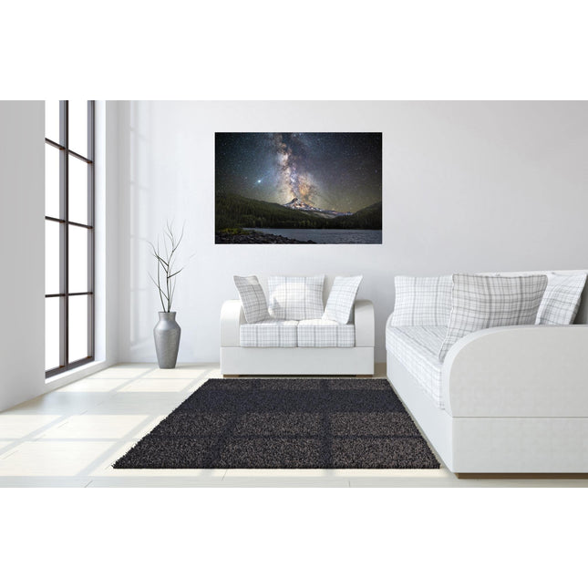A modern living room has white walls, plaid-patterned furniture, and a large dark rug on a light wood floor. A framed Starry Mountain artwork depicting a night sky over forested mountains hangs on the wall. A tall plant in a vase is placed by the window.