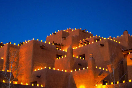 Illuminated by glowing lanterns, the Adobe Sante Fe building shimmers like museum-quality artwork under the deep blue sky. The warm light highlights its terracotta hue, creating a festive atmosphere with bare trees standing elegantly in the foreground.