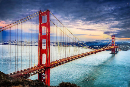 The image, titled Golden Gate, showcases San Franciscos iconic red suspension bridge at sunset. The bridge elegantly stretches over tranquil waters, set against a cloudy sky with the city skyline in the background, creating an art piece of museum quality.