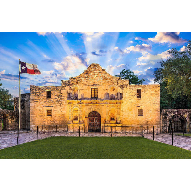 The image depicts the historic stone facade of the Alamo in San Antonio, Texas, at sunset. A vibrant sky lights up museum-quality artwork and a Texas flag waves on the left, framed by a lawn and stone pathway.
