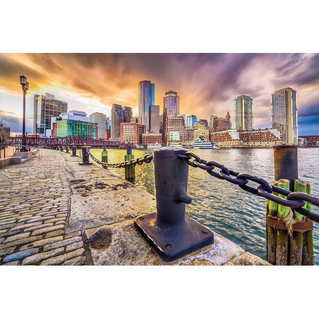 The Boston Harbor captures a city skyline by the water at sunset as museum-quality artwork. Towers reflect the vibrant sky, while a cobblestone path with iron chains borders the waterfront, leading to a bridge beneath stunning orange, purple, and gray clouds in the dramatic sky.