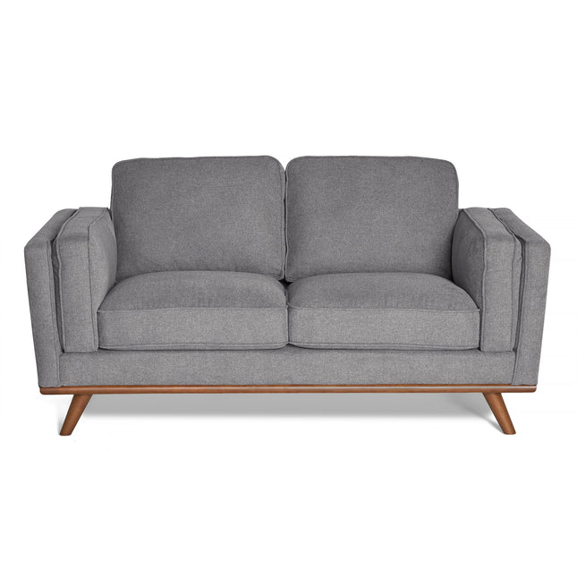 The Maddox Loveseat, a gray, minimalist design with mid-century modern flair, boasts plush cushions, wooden legs, and armrests. Its smooth upholstery complements its durable build, set against a white backdrop.