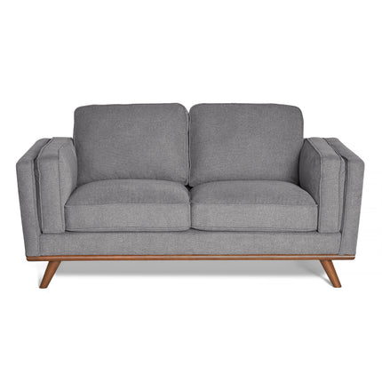 The Maddox Loveseat, a gray, minimalist design with mid-century modern flair, boasts plush cushions, wooden legs, and armrests. Its smooth upholstery complements its durable build, set against a white backdrop.